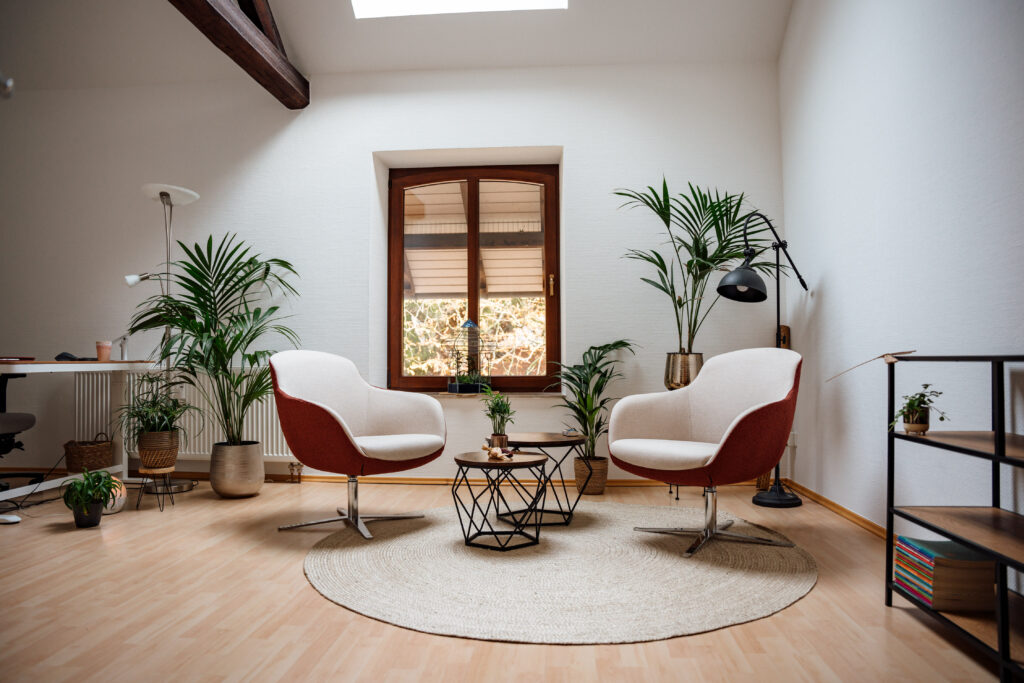 Moderner Therapieraum mit zwei Drehsesseln, Beistelltischen, Lampen, einem Teppich einem Schreibtisch und Zimmerpflanzen.